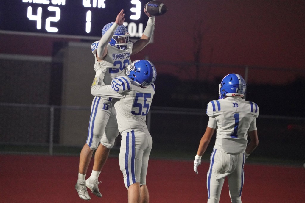 Two football players celebrate a touchdown on the field, with one player jumping in the air while being lifted by another, and a scoreboard visible in the background.