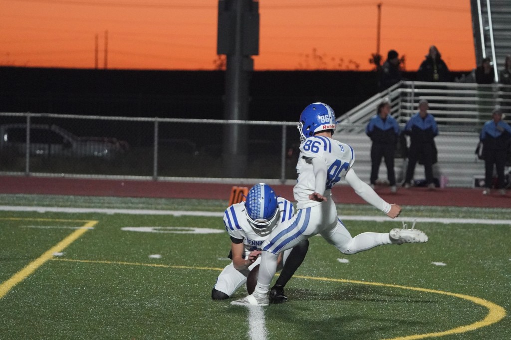 A football player prepares for a kick while another player holds the ball on the field during sunset.