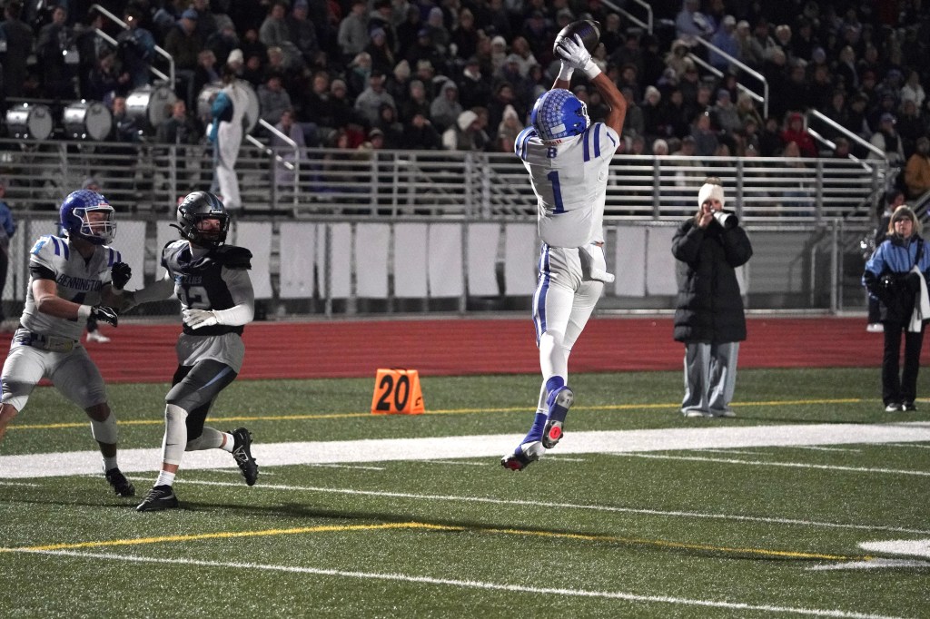 A football player in a white jersey catches a pass during a night game, with opposing players nearby and a crowd in the background.