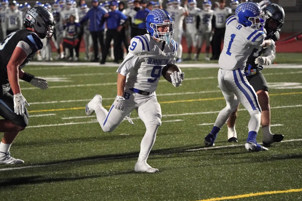 A high school football player in a white jersey with blue accents runs with the ball, avoiding defenders during a night game.