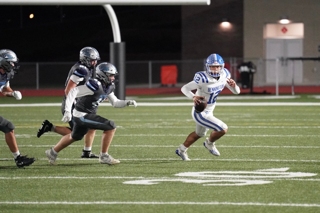A football player wearing a white jersey with blue stripes and the name 'Bennington' runs with the ball while being pursued by two defenders in gray uniforms on a well-lit field.