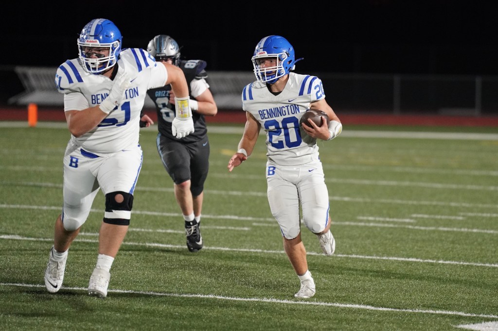 A football player wearing a white and blue uniform with the number 20 runs with the ball on a field, while a teammate in a similar uniform supports him from the left.