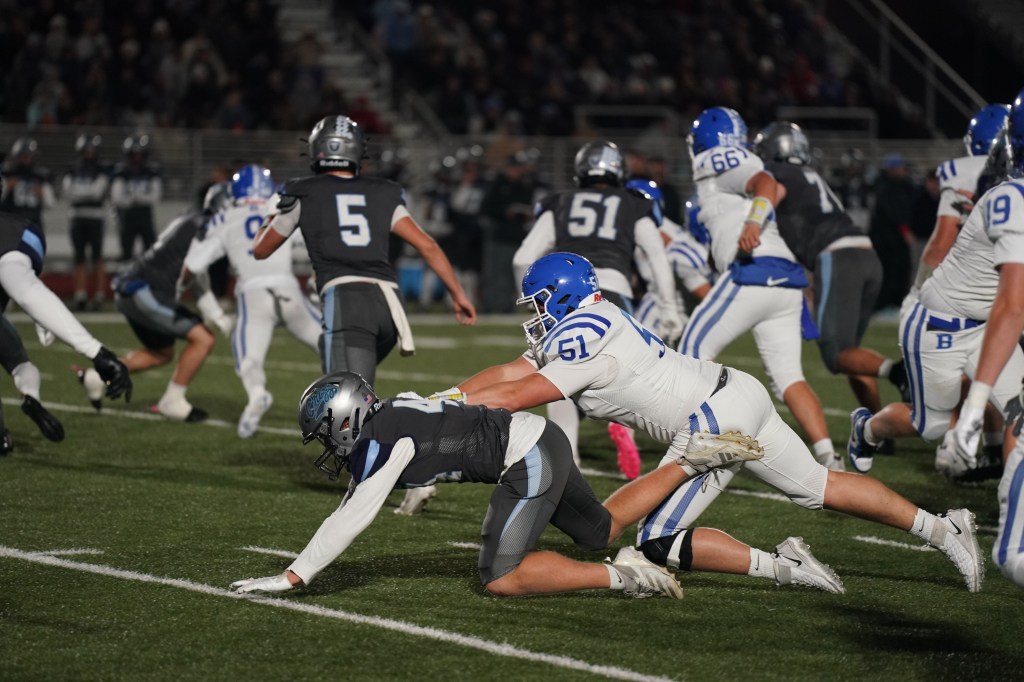 A high school football game in action, featuring players in blue and gray uniforms. One player is tackling another on the field, with a crowd visible in the background.