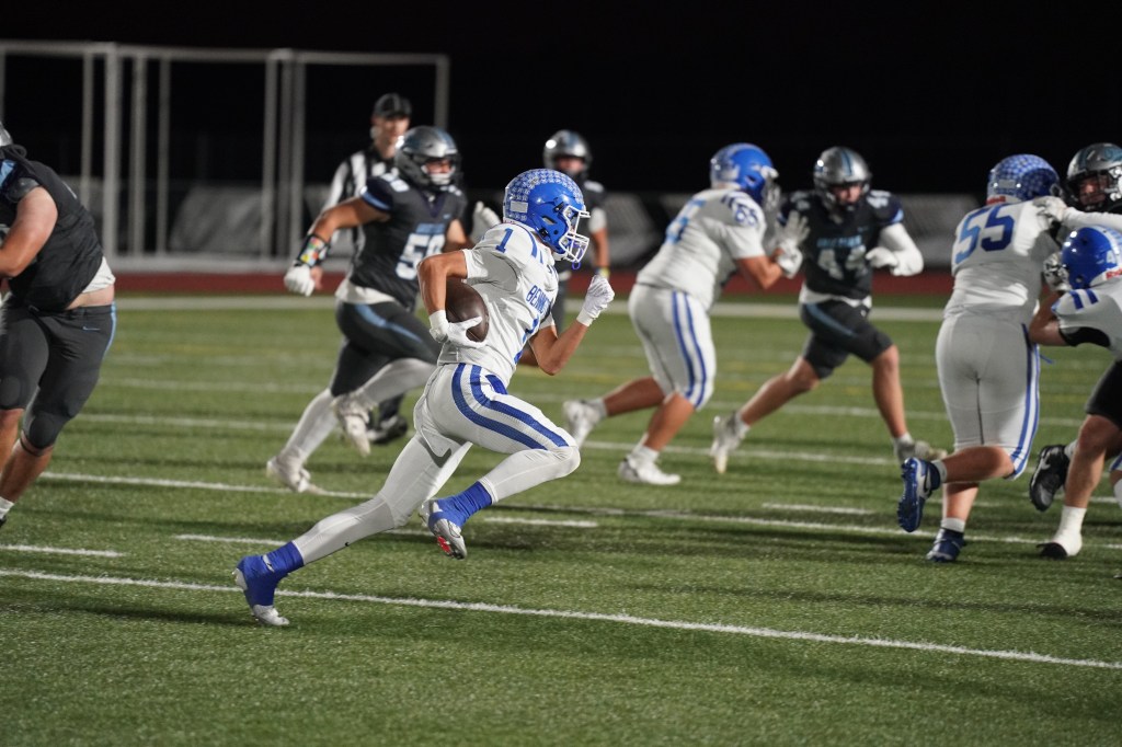 A high school football player in a white and blue uniform runs with the ball on a field, while other players are engaged in a play in the background.