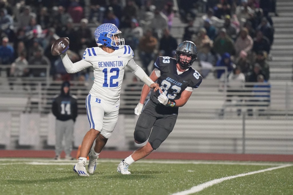 A football player in a white uniform with blue accents, number 12, is throwing a football while being chased by an opponent in a black uniform, number 83, during a night game.