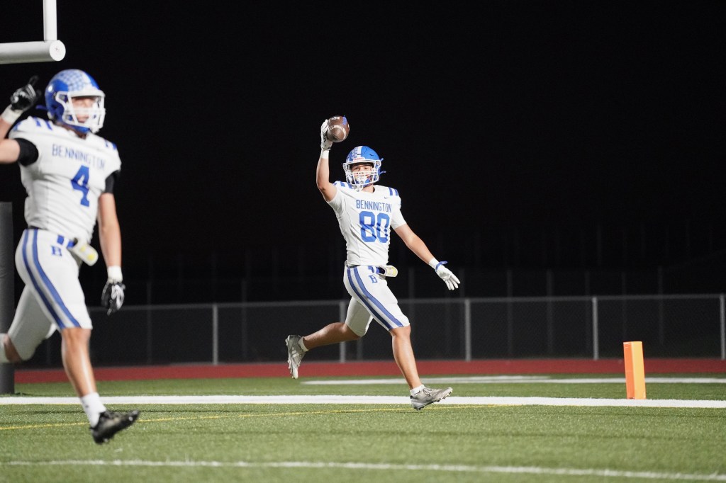 A football player in a white and blue uniform celebrates while holding a football, with a teammate in the background. The scene is set on a football field at night.