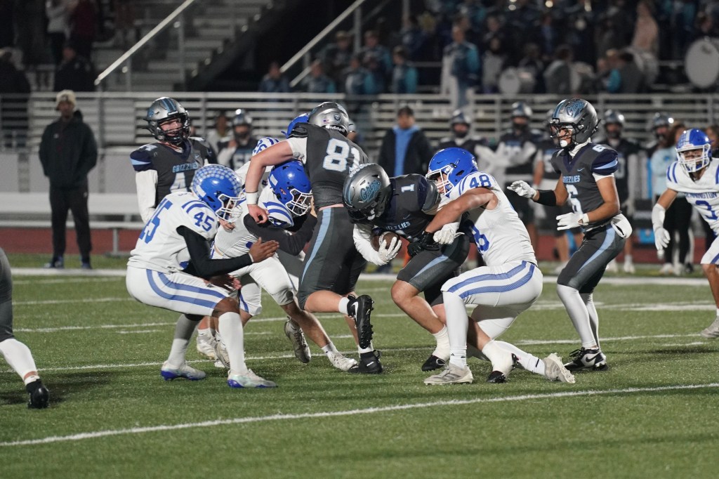 A football game scene with players in action, featuring a player in gray being tackled by two players in blue jerseys.