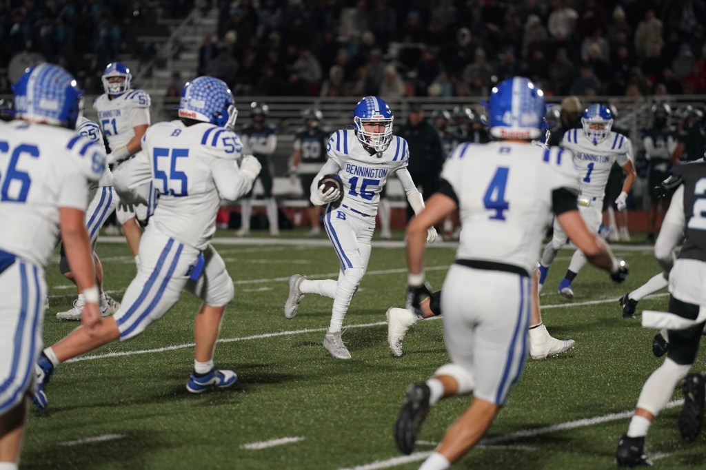 A football player in a white uniform with blue accents runs with the ball, surrounded by teammates and opponents on a nighttime field.