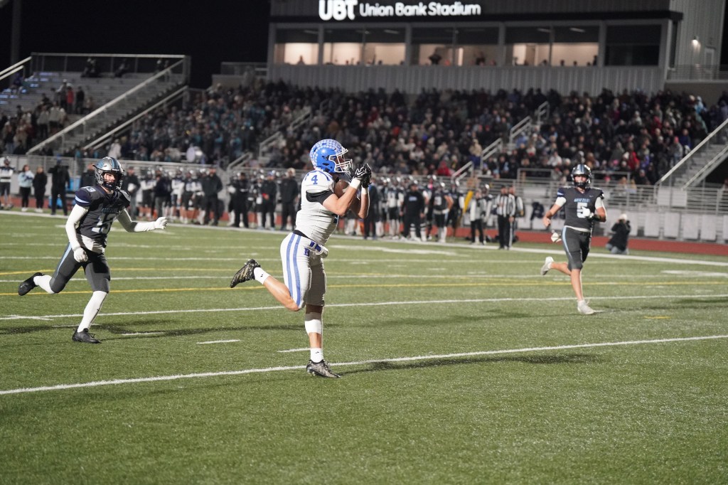 A football player in a gray uniform with blue accents catches a pass on a stadium field, while another player in a black uniform attempts to defend. The stands are filled with spectators in the background.