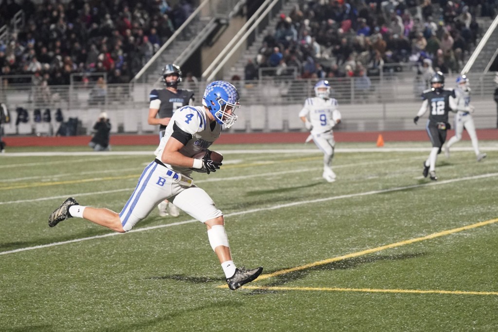A football player in a white uniform with blue accents is running towards the end zone during a night game, with spectators in the stands in the background.