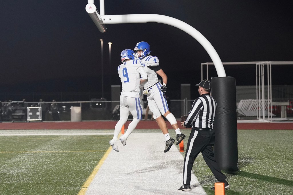 Two football players celebrating after scoring a touchdown under the goalposts at night, with a referee observing.