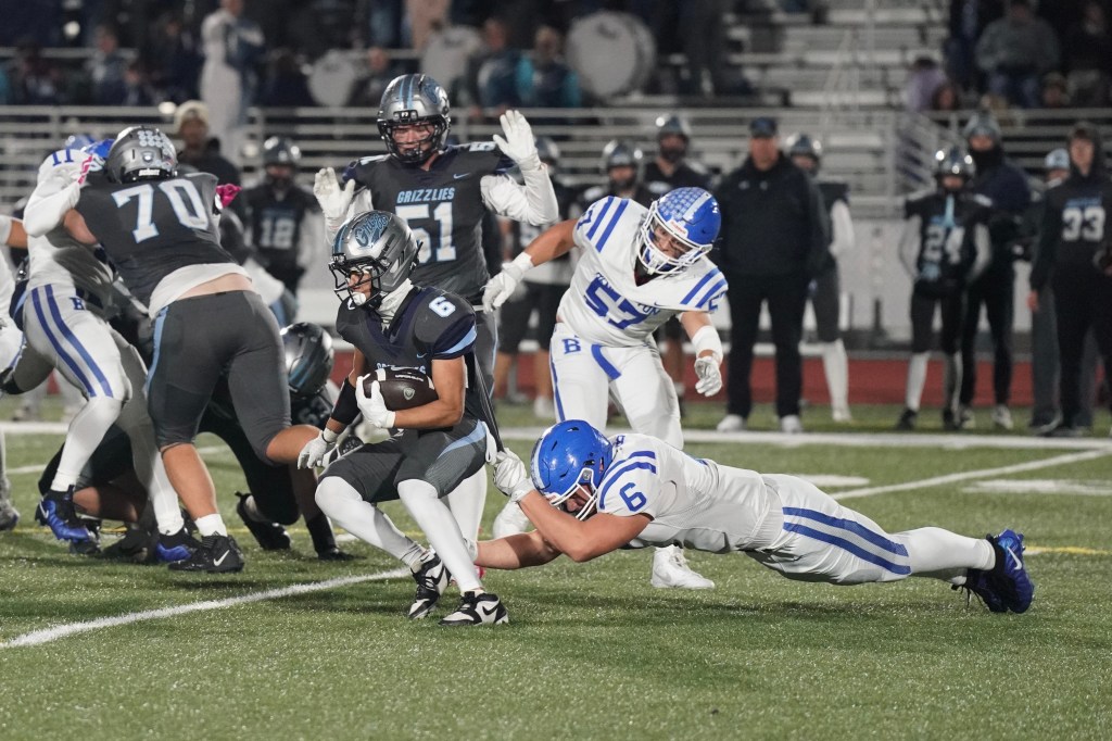 A high school football game in progress, featuring players in blue and gray uniforms. A player wearing number 6 is being tackled by an opponent as teammates surround the action.