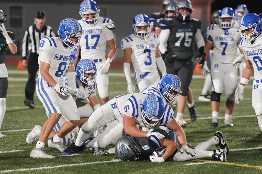 A group of football players in white uniforms with blue accents from Bennington are tackling an opposing player in a dark gray uniform on the field during a game.