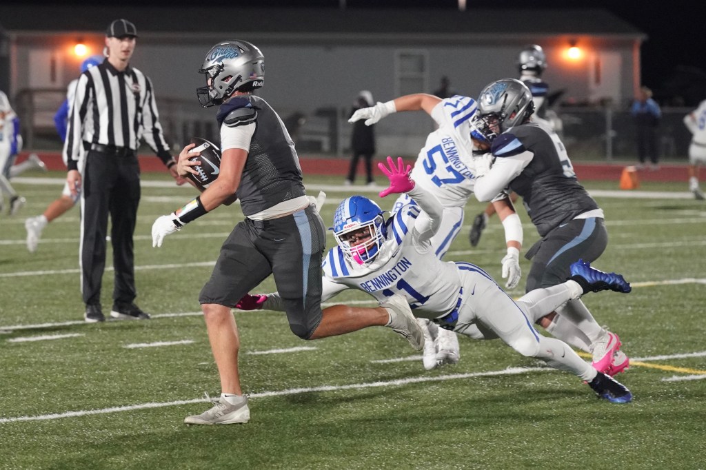 A high school football player in a gray uniform runs with the ball, evading a tackle from an opponent in a white and blue uniform. The referee is visible in the background, observing the play.