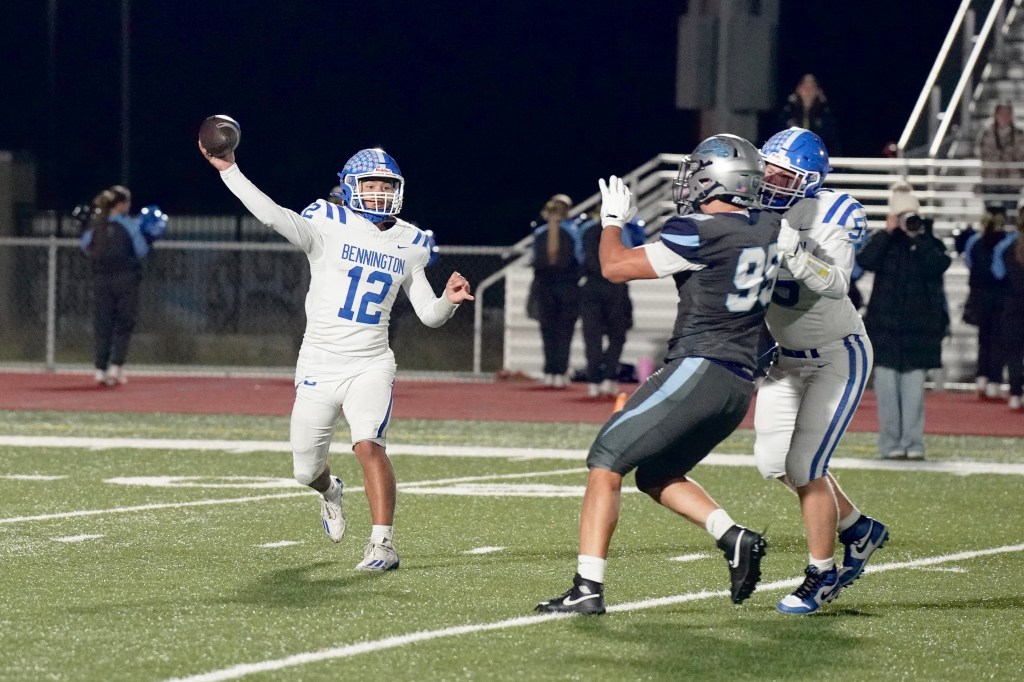 A football player in a white and blue uniform, number 12, is preparing to throw a pass on the field during a night game. He is being pressured by a defender in a gray uniform, number 99, while another teammate offers support.