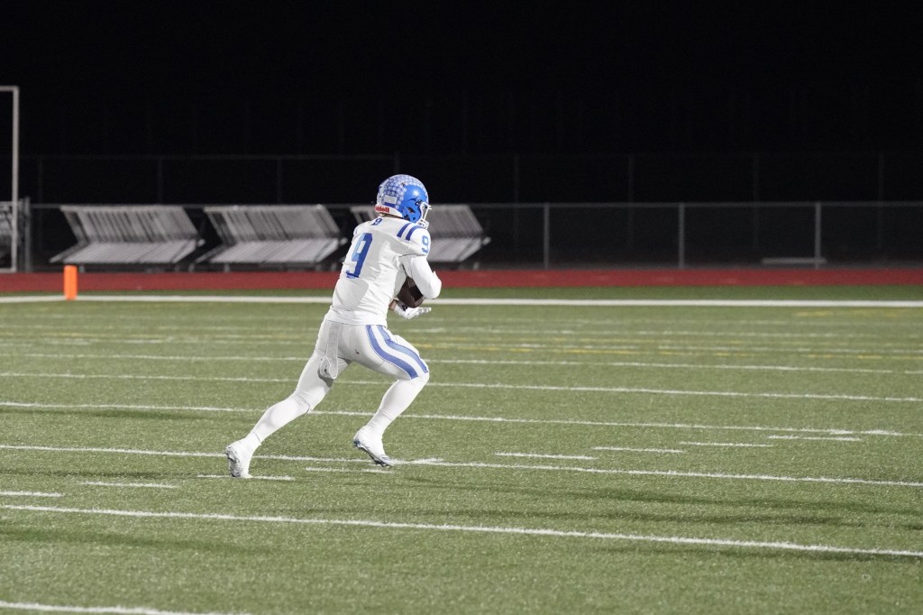 A football player in a white uniform with blue accents running on a field at night, preparing to catch a pass.