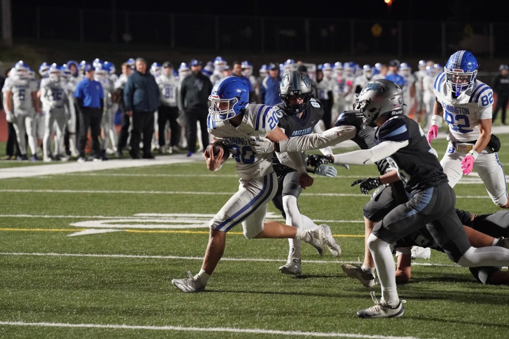 A football player in a blue uniform evades an opponent in a gray uniform while running on the field. In the background, a group of players and spectators watch the action.