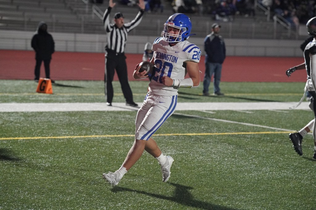 A football player wearing a white jersey with blue stripes, representing Pennington, runs on the field holding a football. In the background, a referee signals, and the stadium is filled with spectators.