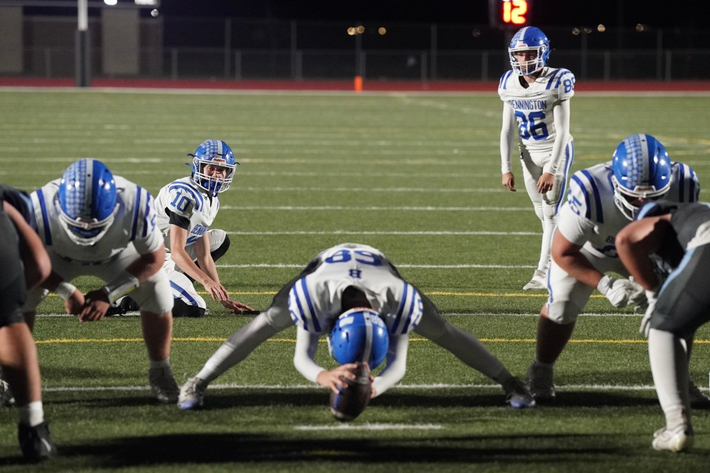 A high school football team preparing for a play on the field at night. Players are in white uniforms with blue accents, huddled around the center who is about to snap the ball.