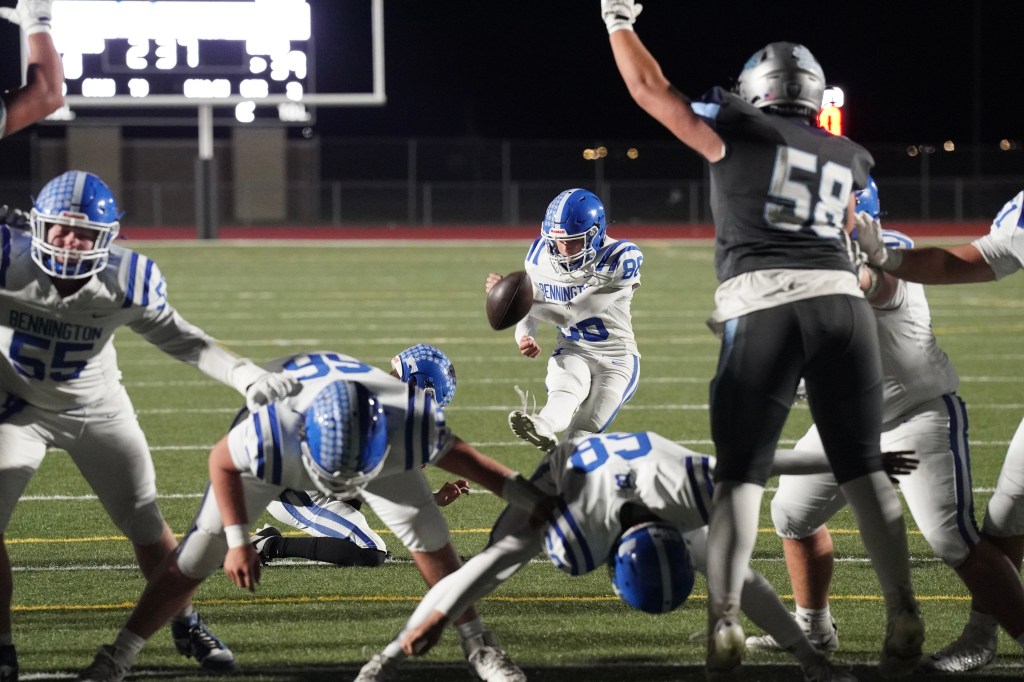 A football player in a blue uniform from Bennington preparing to throw the ball during a night game, with teammates blocking defenders in the foreground.