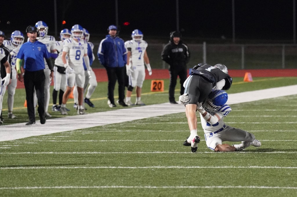 A football player in a gray uniform is being tackled by a player in a blue and white uniform during a game, with teammates watching in the background.