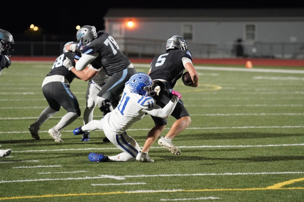 A football player in a black jersey carrying the ball is being tackled by a player in a white and blue uniform on a turf field during a night game.