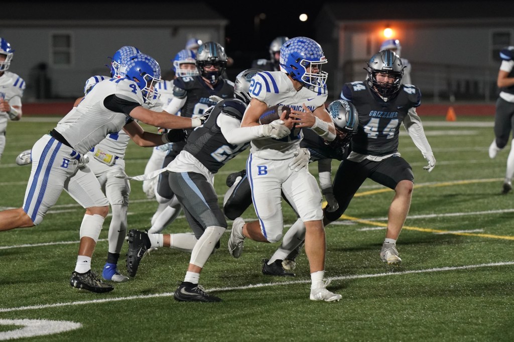 A high school football player in a white uniform is being tackled by defenders in dark gray uniforms during a night game on a well-lit field.