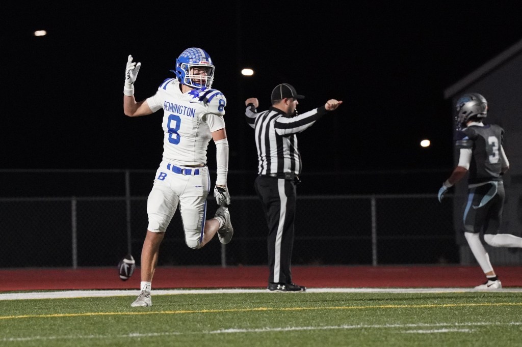 A football player in a white jersey with blue accents celebrates with one hand raised after a play, while a referee signals in the background. Another player in a dark uniform walks away.