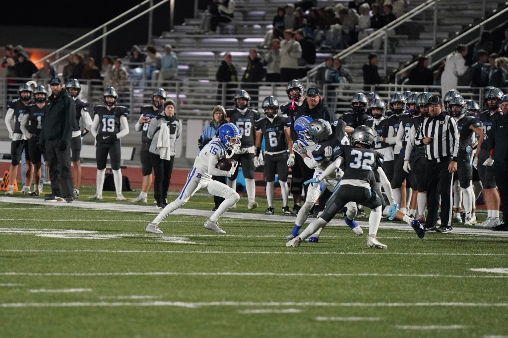 A high school football game in progress, featuring a player in a white uniform with blue accents evading tackle from two players in gray uniforms. The sidelines are filled with players and coaches in the background.
