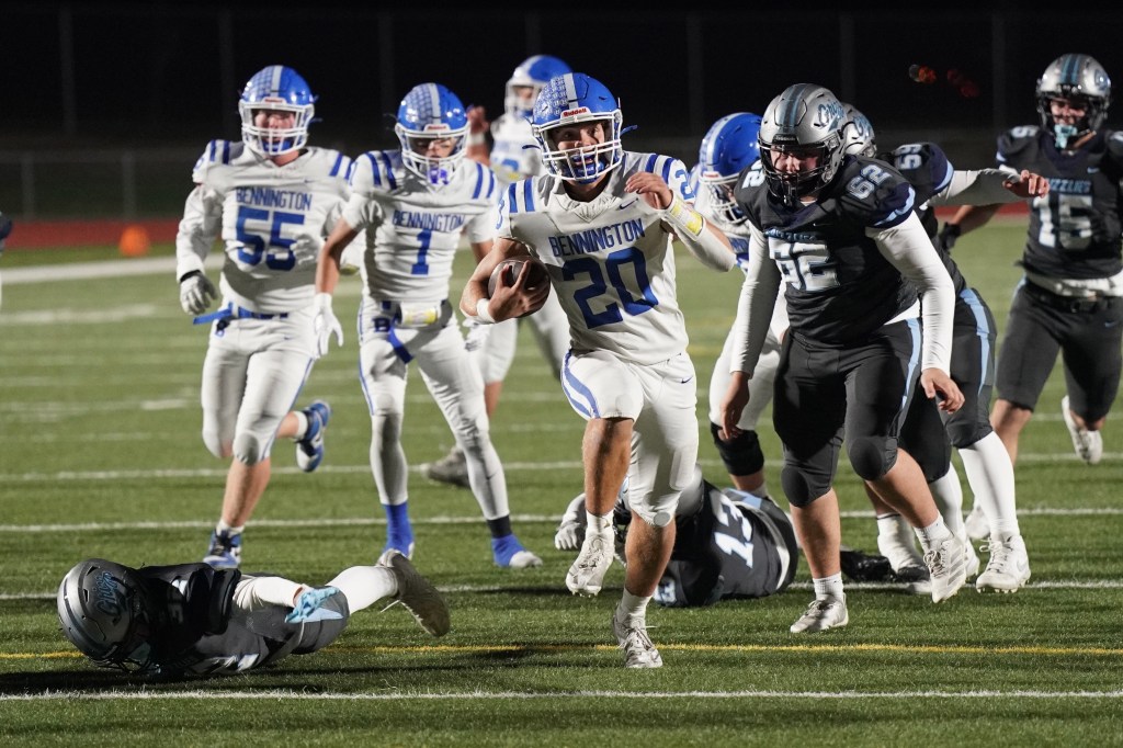 A football player wearing a white and blue uniform runs with the ball during a night game, dodging defenders on the field.