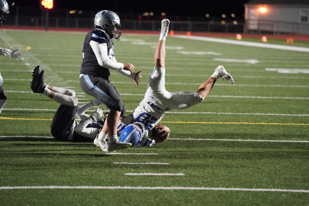 A football player in a blue uniform is falling backward after being tackled by another player in a black uniform on a sports field at night.
