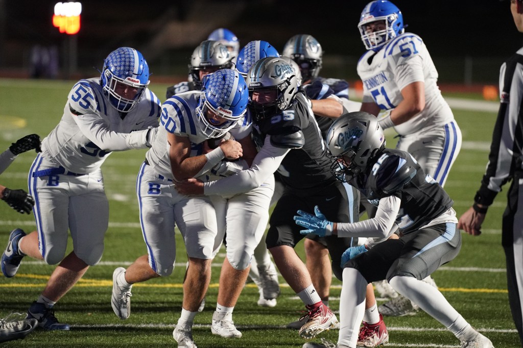 A high school football game with players from two teams in action; one team in blue and white uniforms is attempting to tackle a player in a black uniform, while other players are engaged nearby.