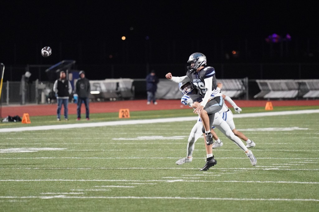A football player throwing a football during a night game, with two opposing players in the background.