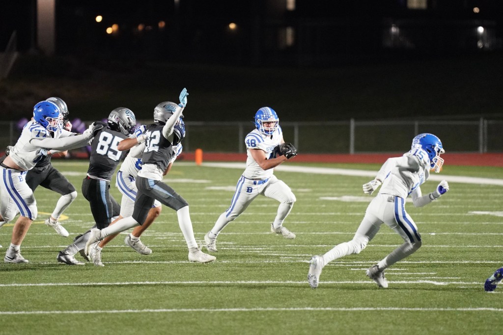 A football player in a white uniform with blue accents runs with the ball, evading defenders on a field at night.