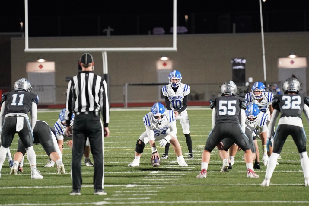 A football game in progress with players from two teams lined up on the field. A referee is overseeing the action as the offense prepares to snap the ball.