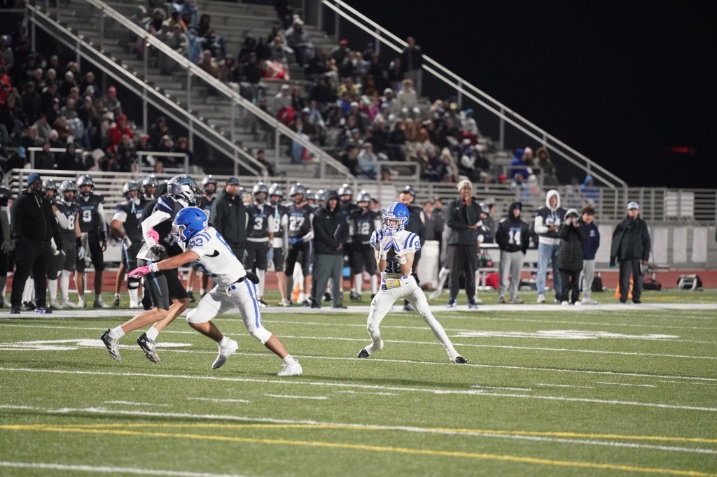 A high school football game in progress at night, featuring players in blue and white uniforms. One player is catching a pass while another is attempting to tackle an opponent. The stands in the background are filled with spectators.