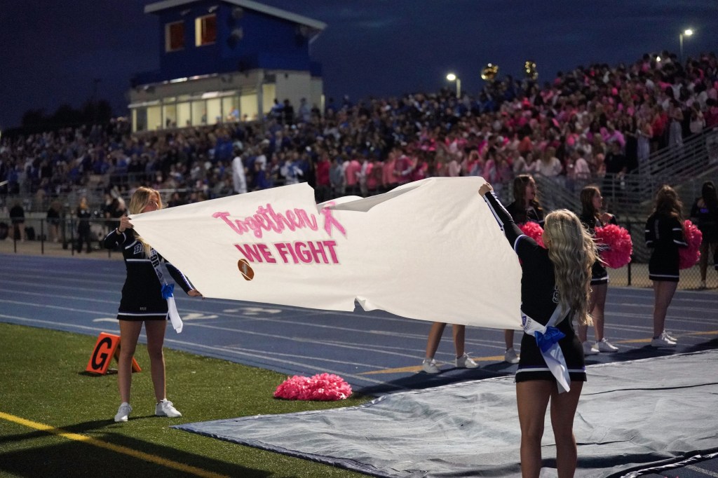 Cheerleaders holding a banner that reads 'Together We Fight' at a nighttime sports event, with a crowd in the background.
