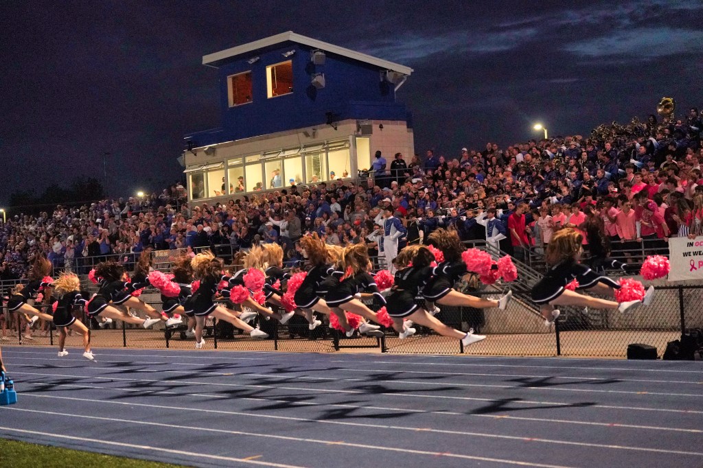 A group of cheerleaders performing a jump with pom-poms during a night sports event, with a large crowd in the background.