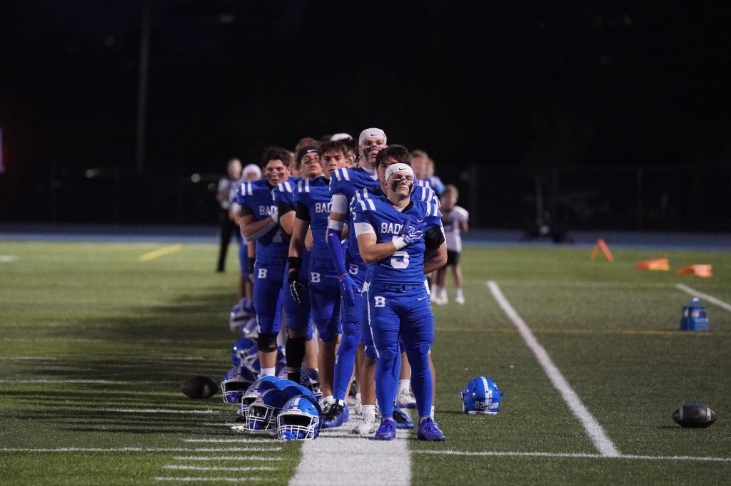 A lineup of high school football players wearing blue uniforms, standing on the field at night.
