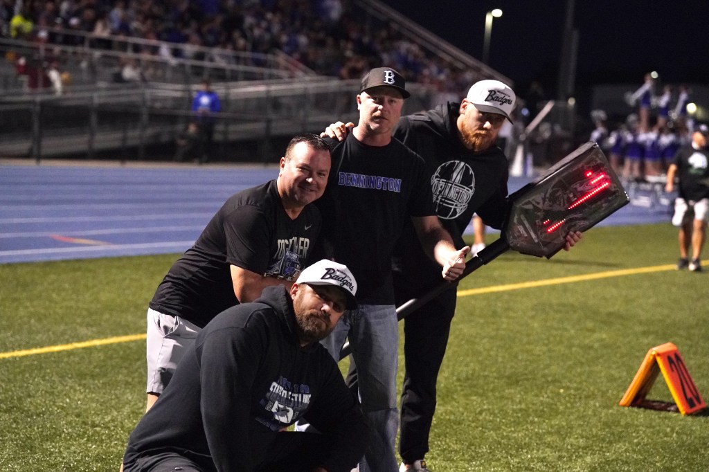 A group of four coaches or team members on the sidelines of a football game, posing playfully with sports equipment in a nighttime setting.