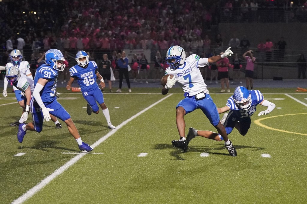 A high school football player wearing a white jersey with blue accents dodges a tackle on a grassy field, while teammates and opponents are in motion around him.