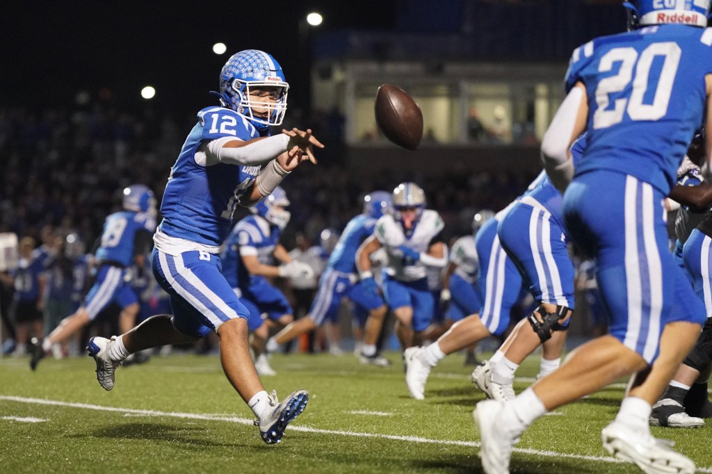 A high school football player in a blue uniform with the number 12 is throwing a football, with several teammates running in the background during a night game.