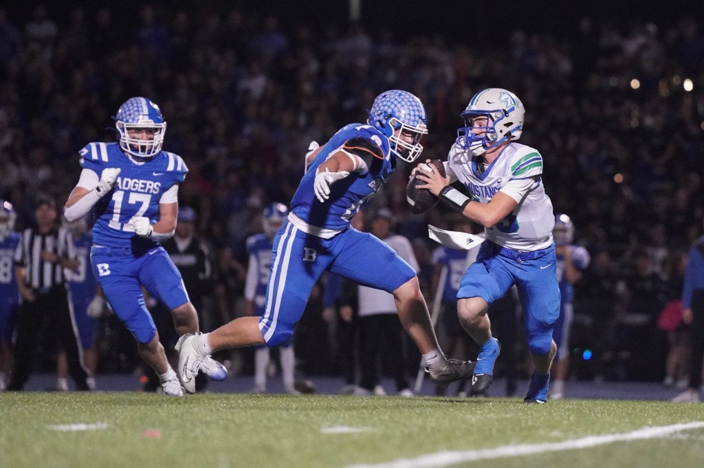 A high school football player in blue uniform is tackling an opponent in white uniform during a game, with spectators in the background.