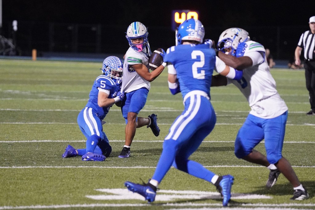 A high school football player wearing a white jersey runs the ball while being pursued by defenders in blue jerseys on a football field.