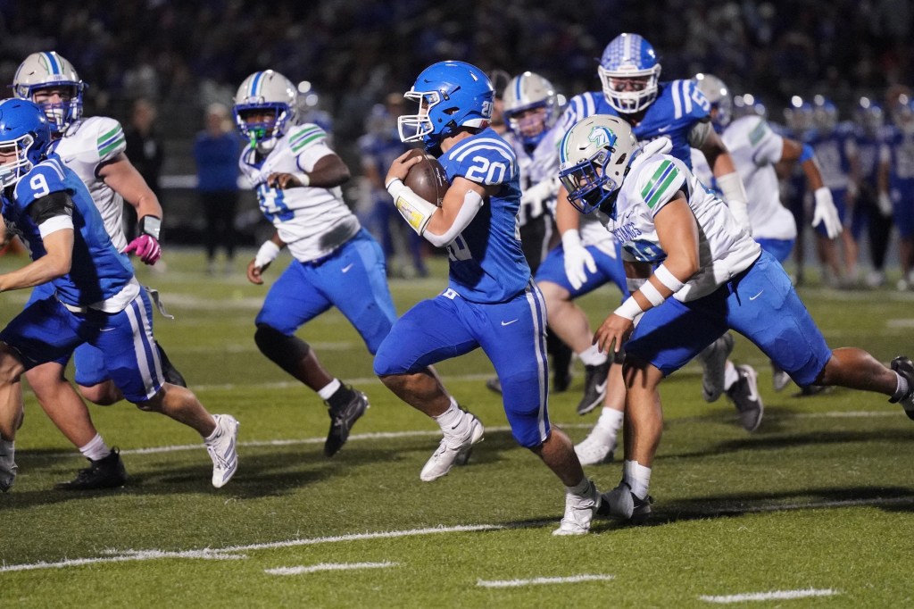 A high school football game in action, featuring players in blue and white uniforms. A player wearing number 20 is running with the ball while being pursued by an opponent from the opposing team.