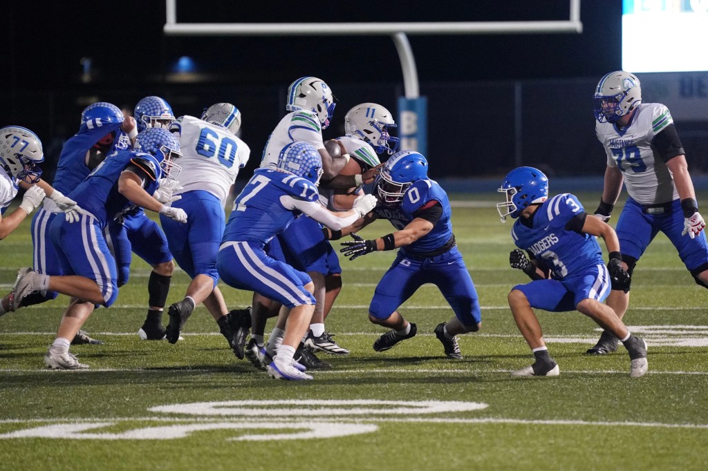 A dynamic scene of a high school football game, showing players in blue uniforms on the field. One player, number 0, is attempting to tackle an opposing player, while teammates support from the side.