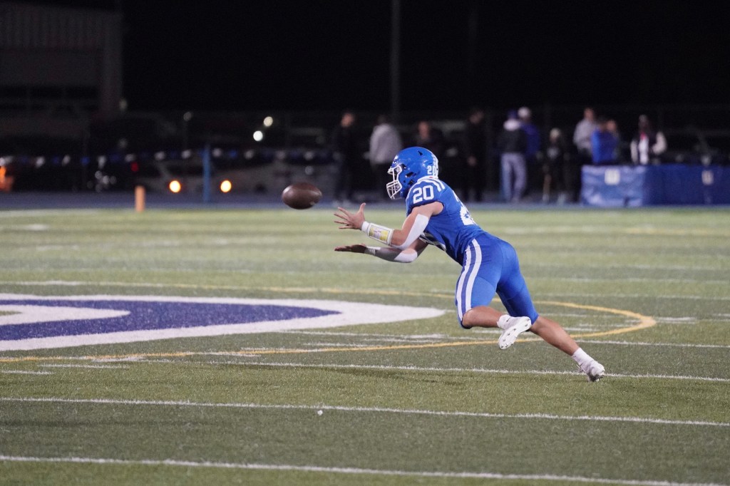 A football player in blue jersey number 20 dives to catch a football on a field at night.