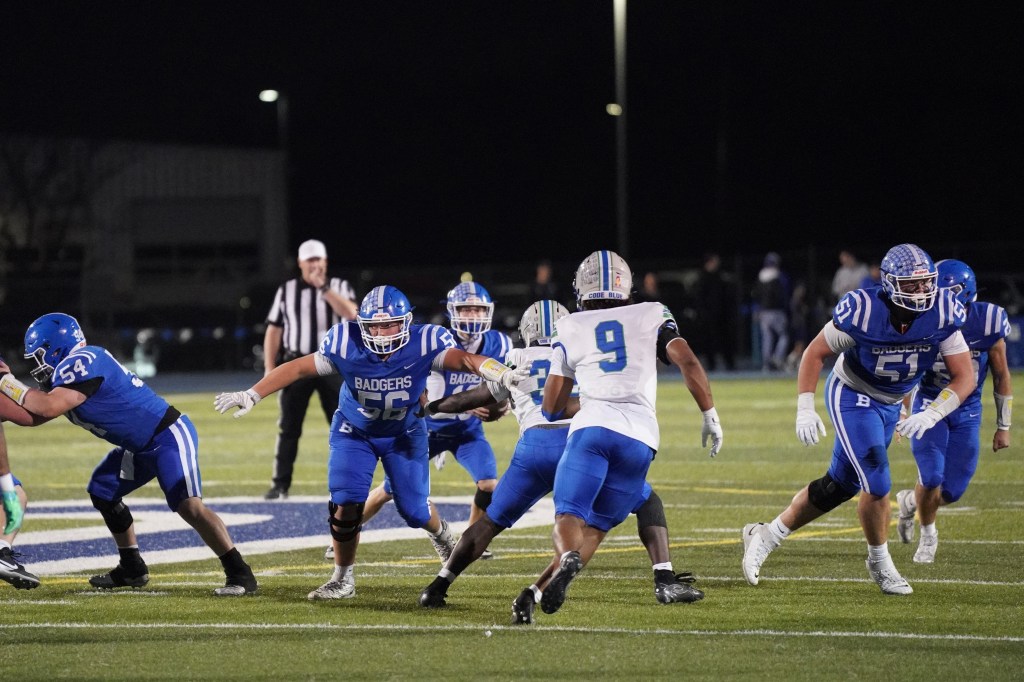 An intense high school football game in progress, with players in blue uniforms (representing the Badgers) on defense, while an opposing player in white attempts to advance the ball.