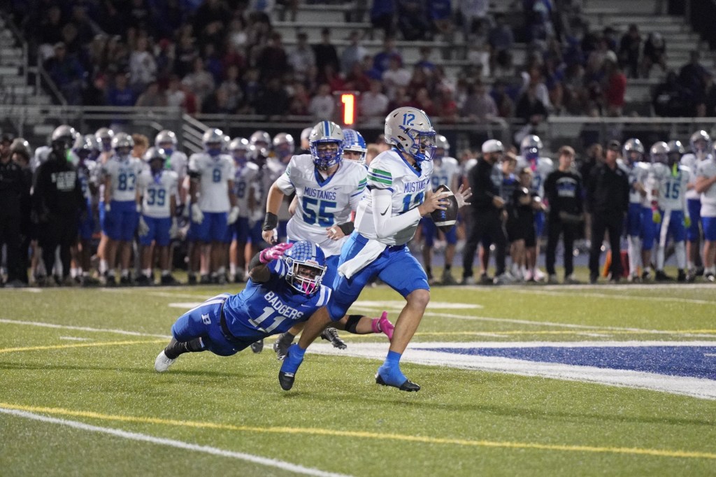 A high school football player in a blue uniform running with the ball while avoiding a tackle from a player in a blue and white uniform. A crowd of spectators is visible in the background.
