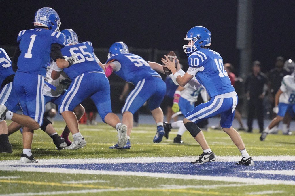 A high school football player in a blue jersey number 10 prepares to throw a football while being defended by players on the field.